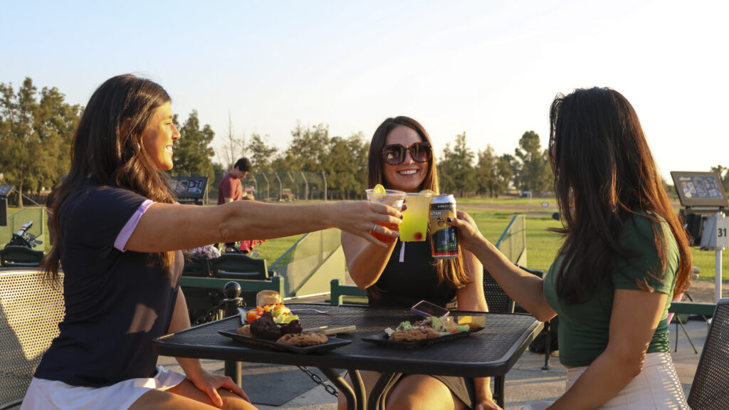 Three women enjoying drinks and appetizers at a golf driving range, toasting at an outdoor table overlooking the practice area during a social golf outing.