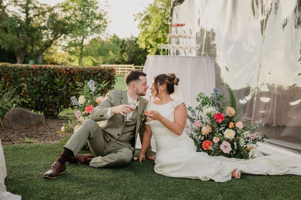 Bride and groom sharing a toast beside floral arrangements outside a reception tent at The Pavilion at Haggin Oaks in Sacramento.