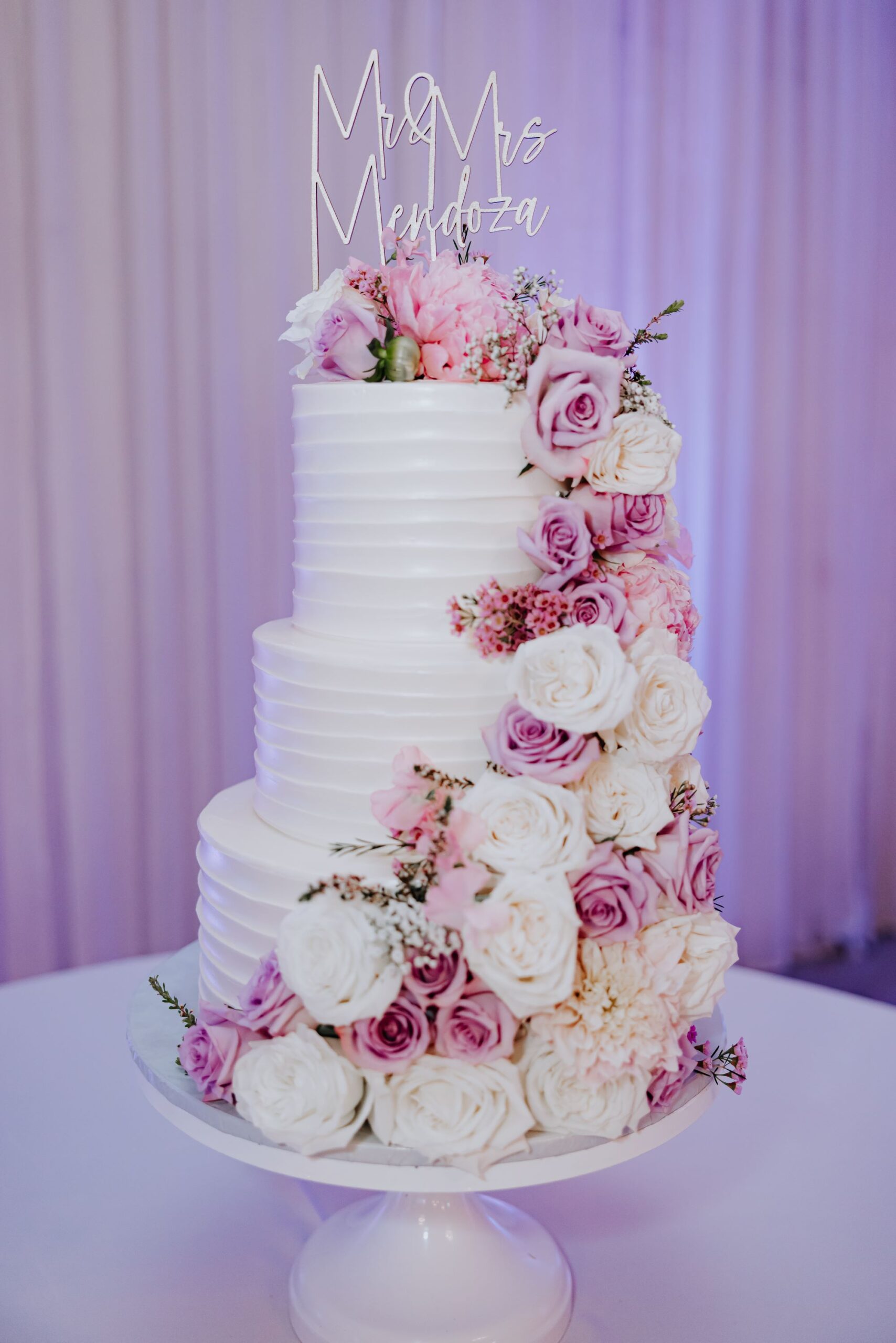 Wedding cake with cascading pink and white floral arrangements and a personalized cake topper displayed at a reception inside The Pavilion at Haggin Oaks in Sacramento.