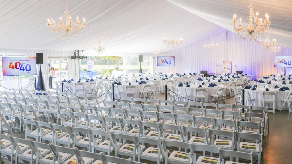 Corporate event setup inside a large white reception tent at The Pavilion at Haggin Oaks featuring chandeliers, banquet tables, presentation screens, and theater-style seating in Sacramento.