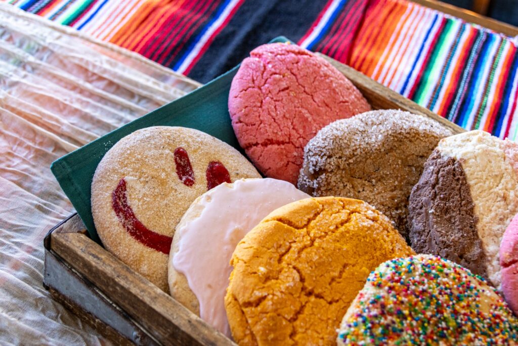 Traditional Mexican pan dulce assortment displayed on a colorful serape tablecloth, featuring conchas, sugar cookies, and assorted sweet breads for a cultural celebration or catered event.