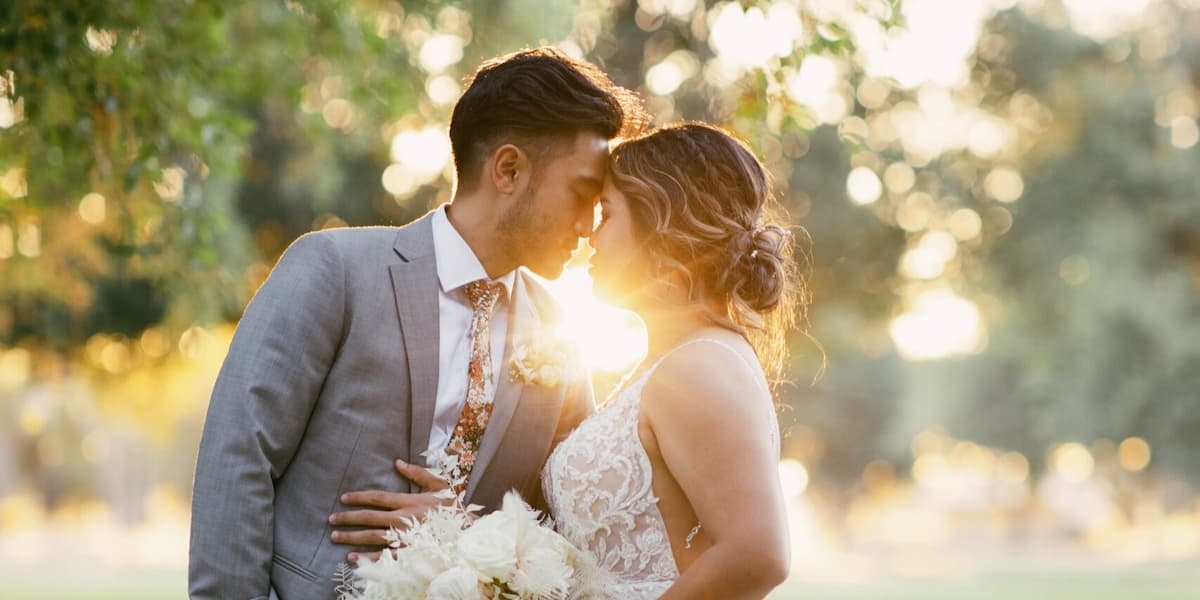 Romantic outdoor wedding portrait of a bride and groom embracing at golden hour, with soft sunlight glowing between them and blurred greenery in the background.