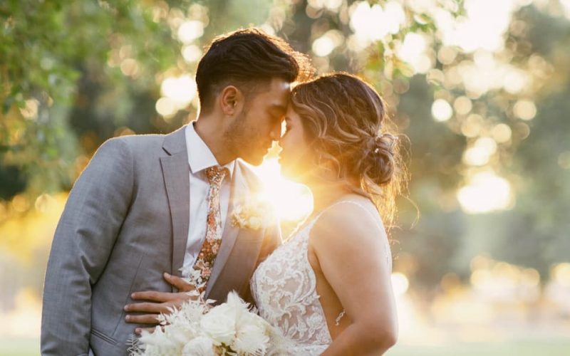 Romantic outdoor wedding portrait of a bride and groom embracing at golden hour, with soft sunlight glowing between them and blurred greenery in the background.